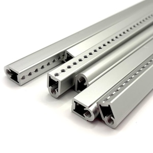 May include: Four silver-colored metal extrusions with a smooth finish. Each piece features a linear row of small, evenly spaced circular holes along one edge and a hollow, shaped channel running through the center of the profile.