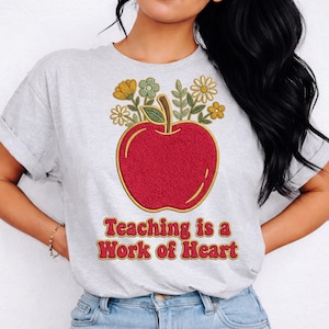 Puede incluir: Camiseta gris claro con una gran manzana roja con contorno dorado y un ramo de flores. El texto "Teaching is a Work of Heart" está impreso debajo en una fuente retro.