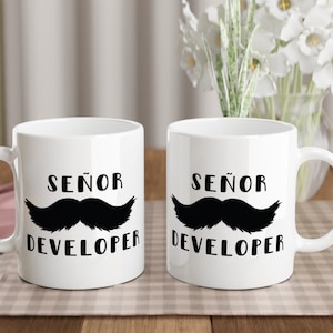 Puede incluir: Dos tazas de café blancas con las palabras "SEÑOR DEVELOPER" y un gráfico de bigote negro. Las tazas están sobre un mantel a cuadros con utensilios de madera y un paño rosa. Un jarrón con flores blancas está en el fondo.