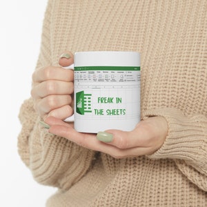 Può includere: Tazza in ceramica bianca con un design a foglio di calcolo e la scritta "FREAK IN THE SHEETS" in verde. La tazza ha un manico ed è tenuta da una persona. La tazza ha un'icona a forma di grafico verde.