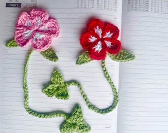 Puede incluir: Dos marcapáginas de flores de ganchillo con hojas y tallos verdes. Las flores son rosas y rojas con centros blancos.