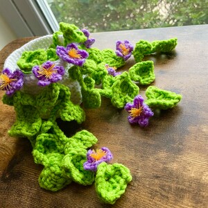 May include: A crocheted plant with green leaves and purple flowers in a white pot. The plant is hanging over the edge of a wooden table.