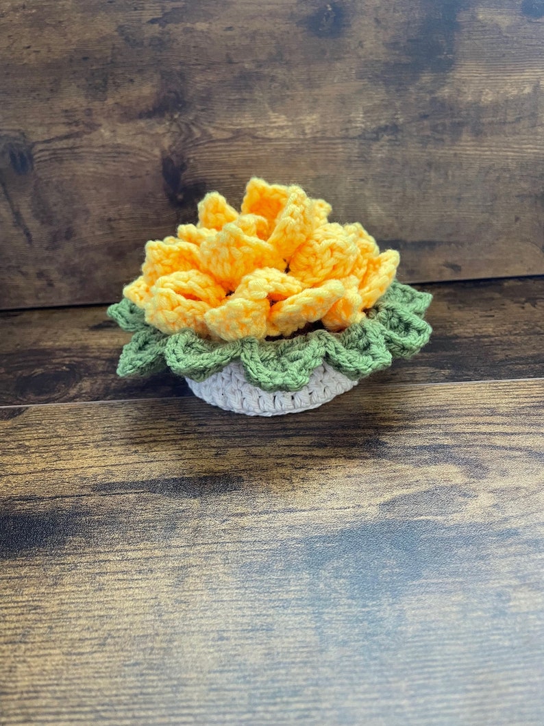 May include: A crocheted flower in a yellow and green pot. The flower is made of yellow yarn and the pot is made of green and white yarn.