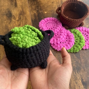 May include: A black crocheted cauldron filled with green crocheted leaves, surrounded by pink and green crocheted coasters.
