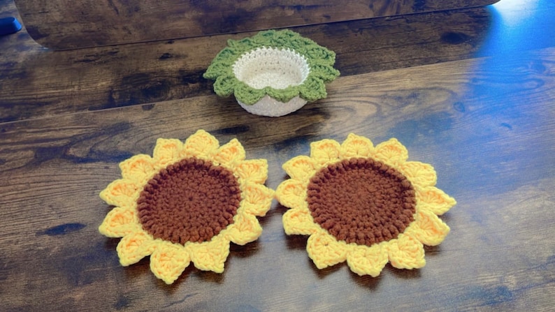 May include: Two crocheted sunflower coasters with brown centers and yellow petals. A crocheted green and white bowl is also in the image.