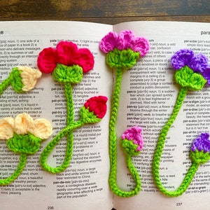May include: Four crocheted bookmarks with flower designs. The bookmarks are in pink, yellow, purple, and green. The bookmarks are on a page of a dictionary with the words "page", "pale", "panel", "parallelogram", and "par-a-ble" visible.