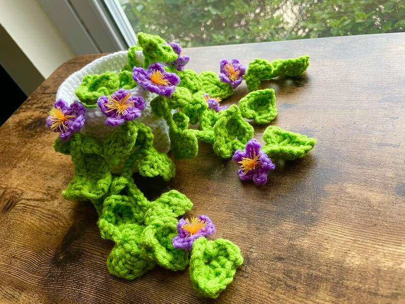 May include: A crocheted plant with green leaves and purple flowers in a white pot. The plant is hanging over the edge of a wooden table.