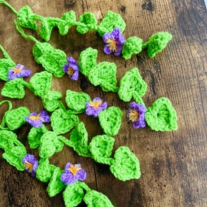 May include: A garland of crocheted green leaves and purple flowers. The garland is made of yarn and is perfect for adding a touch of nature to your home decor.