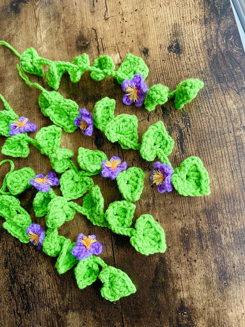 May include: A garland of crocheted green leaves and purple flowers. The garland is made of yarn and is perfect for adding a touch of nature to your home decor.