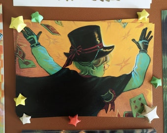 May include: Illustration of a magician with a black top hat, cape, and gloves, surrounded by playing cards. The artwork features a yellow and orange background with star accents. Origami stars of various colors are placed around the illustration.
