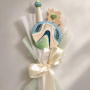 May include: A decorative bouquet featuring a tall tower ornament, a circular plaque depicting the Azadi Tower in blue and cream, and a wooden piece with a green heart. The arrangement is wrapped in translucent white paper with a cream ribbon.