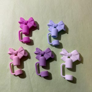 May include: Five silicone pencil toppers shaped like bows in different colors: pink, purple, light blue, white, and lavender. Each topper has a small bell-shaped charm and a loop for attaching to a pencil.