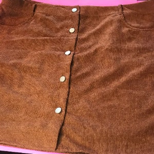 May include: A brown corduroy skirt with a button-up front and two pockets. The skirt is a mini length and has a slightly A-line shape.