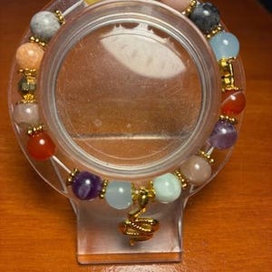 Pu&ograve; includere: Un braccialetto di perline con una variet&agrave; di pietre preziose colorate, tra cui gialle, rosa e blu. Il braccialetto &egrave; impreziosito da perline color oro e un ciondolo dorato. Viene esposto su un supporto di plastica trasparente.