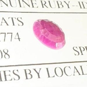 Könnte beinhalten: Nahaufnahme eines rosafarbenen Rubin-Edelsteins. Der Rubin ist facettiert und hat eine glänzende, polierte Oberfläche. Der Rubin ist vor einem weißen Hintergrund mit schwarzem Text platziert, der "GENUINE RUBY - ID ATS 774 08 IES BY LOCAL SP" lautet.
