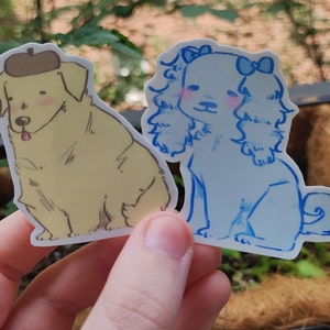 May include: Two illustrated dog stickers. One is a yellow dog wearing a brown hat. The other is a blue dog with a blue bow.