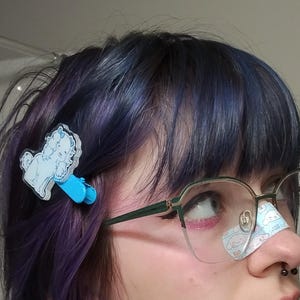 May include: A person with blue and purple hair is wearing glasses and a blue and white dog hair clip. The person has a small bandage on their nose with a cartoon dog on it.