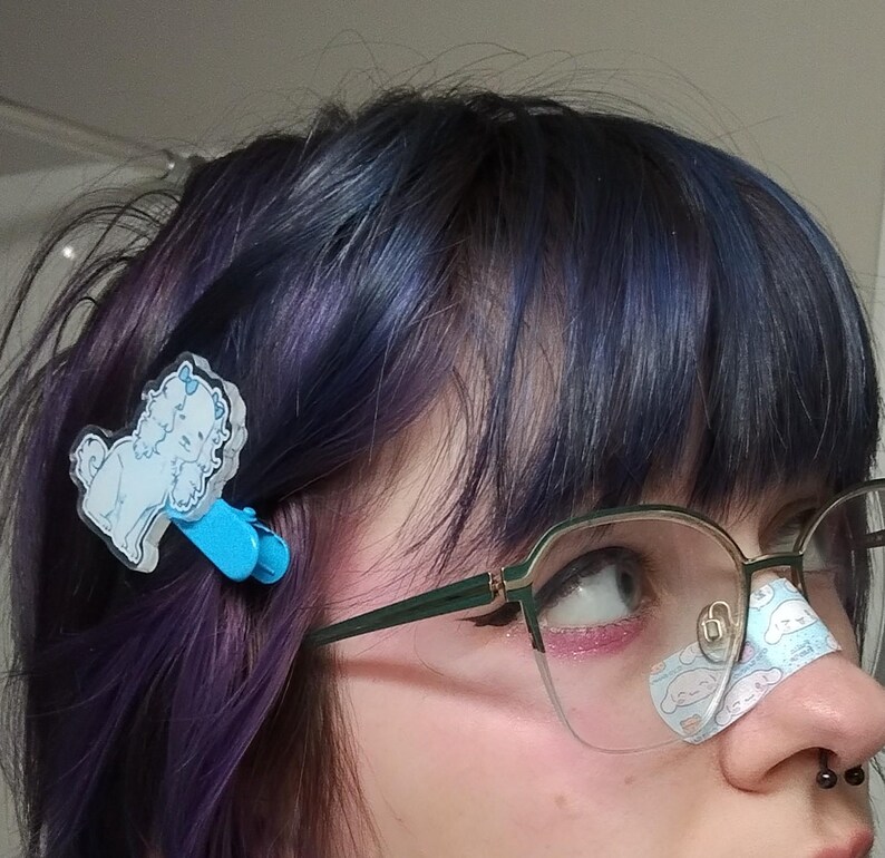 May include: A person with blue and purple hair is wearing glasses and a blue and white dog hair clip. The person has a small bandage on their nose with a cartoon dog on it.