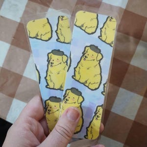 May include: Two clear plastic bookmarks with a repeating pattern of yellow dogs wearing black berets on a light blue background.