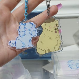 May include: Two clear acrylic keychains with dog illustrations. The keychain on the left features a white dog with a pink bow. The keychain on the right features a yellow dog wearing a brown beret.