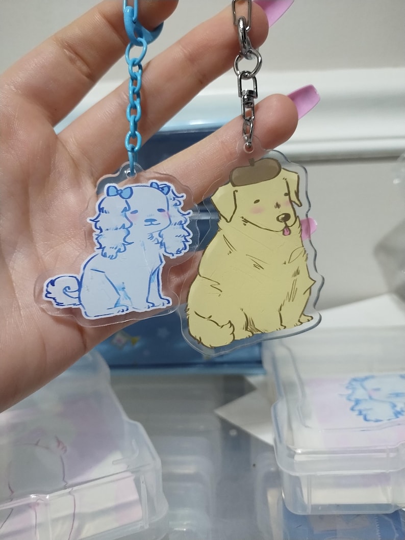 May include: Two clear acrylic keychains with dog illustrations. The keychain on the left features a white dog with a pink bow. The keychain on the right features a yellow dog wearing a brown beret.