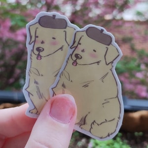 May include: Two yellow dog stickers with brown berets. The stickers are drawn in a cartoon style and have a simple, whimsical look.