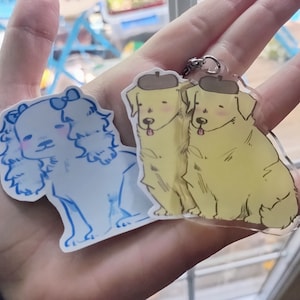 May include: Two dog charm keychains. One keychain features a white dog with floppy ears and a blue bow. The other keychain features two yellow dogs wearing hats.