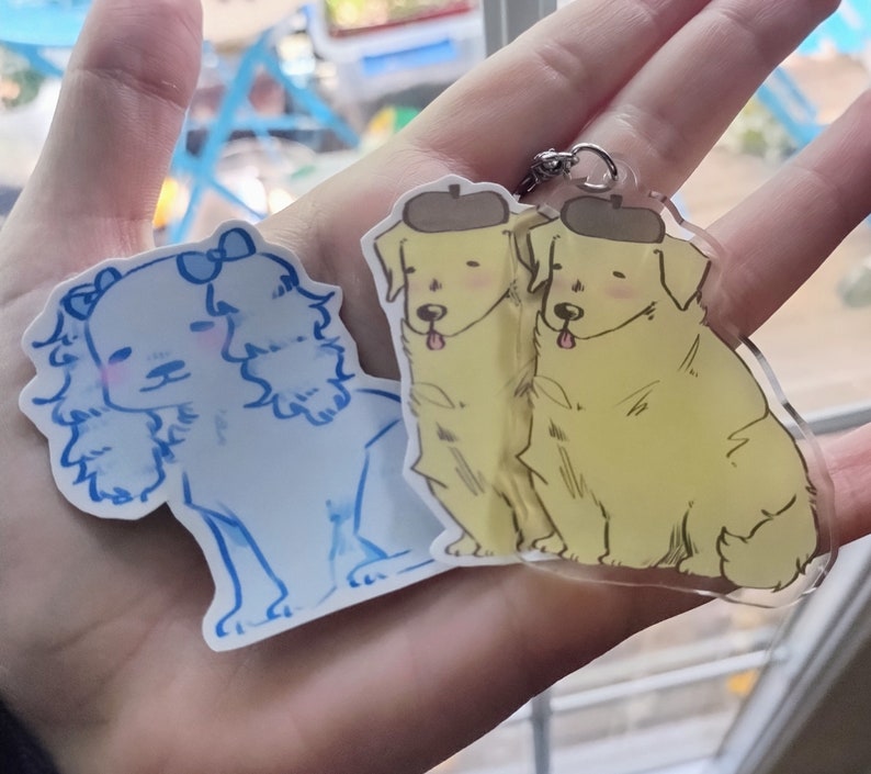 May include: Two dog charm keychains. One keychain features a white dog with floppy ears and a blue bow. The other keychain features two yellow dogs wearing hats.