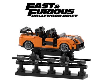 May include: An orange toy car with black racing stripes and a black undercarriage, built from interlocking blocks. The car is mounted on a black track system. The words "FAST & FURIOUS HOLLYWOOD DRIFT" are printed above the car.