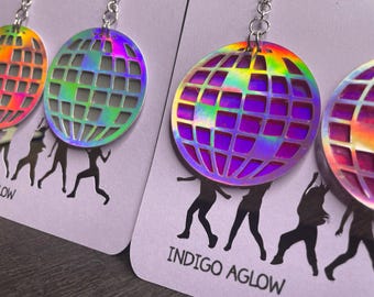 May include: Holographic disco ball earrings with a grid pattern, hanging from silver-colored hooks. The earrings are displayed on a light purple card with the text "Indigo Aglow" and a silhouette of dancing figures.