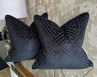Op de afbeelding: Twee zwarte fluwelen kussens met een geometrisch patroon. De kussens liggen op een glazen tafel met een gouden basis.