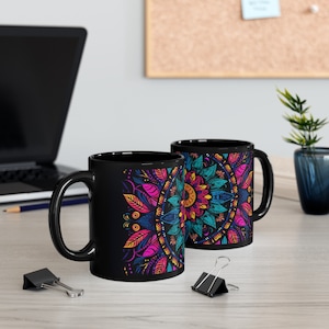 May include: Two black ceramic mugs with a vibrant, colorful mandala design. The mandala features intricate patterns in shades of pink, blue, yellow, and green. The mugs are on a light-colored wooden surface, with a laptop and office supplies in the background.