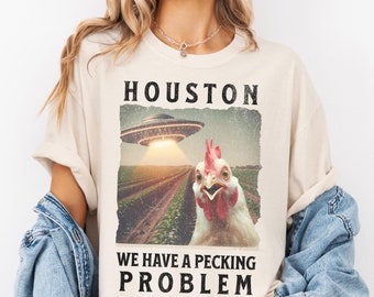 May include: An ivory t-shirt featuring a graphic of a chicken in a field with a UFO hovering above. The text HOUSTON WE HAVE A PECKING PROBLEM is printed in black block letters above and below the image. The shirt has short sleeves and a crew neck.