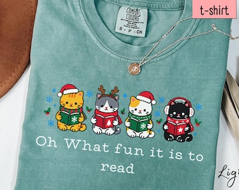 May include: A light green t-shirt featuring a graphic of four cats wearing holiday accessories and reading books. The text "Oh What fun it is to read" is printed below the cats. The shirt has a Comfort Colors label at the neck.