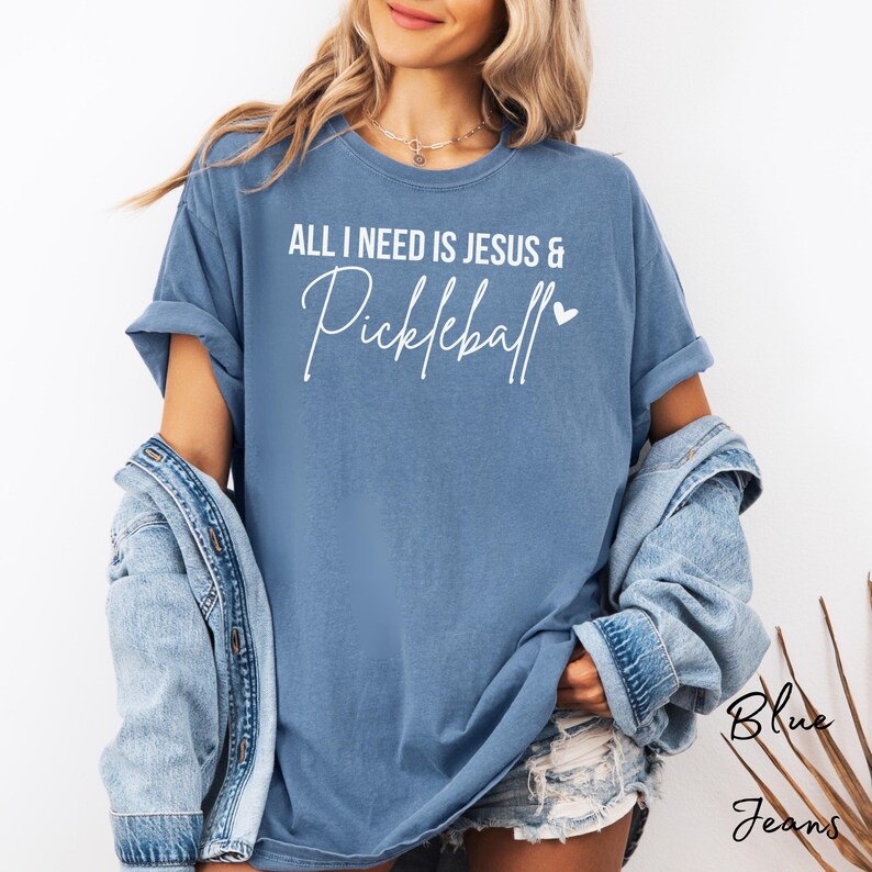 Puede incluir: Una camiseta azul con el texto "All I need is Jesus & Pickleball" en letras blancas con un peque&ntilde;o coraz&oacute;n.