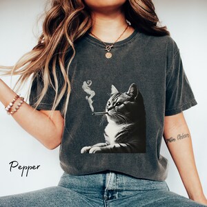 Pu&ograve; includere: Una t-shirt a maniche corte color grigio antracite con una stampa grafica in bianco e nero di un gatto con una sigaretta e fumo che sale. La maglietta ha un girocollo e una finitura lavata. La parola Pepper &egrave; stampata in nero.