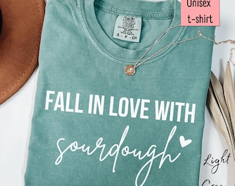 以下が含まれることがあります： FALL IN LOVE WITH sourdoughという白い文字と小さなハートのアイコンがプリントされた、ライトグリーンの半袖Tシャツ。内側の襟元にはComfort Colorsのブランドラベルがあり、ゴールドトーンのペンダントネックレスが添えられています。