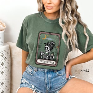 May include: A green t-shirt featuring a graphic of a skeleton holding a cup of coffee. The graphic is in the style of a tarot card and reads "XVI The Coffee Lover".