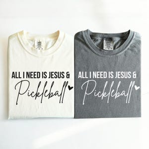Puede incluir: Dos camisetas con el texto "All I need is Jesus & Pickleball" impreso en ellas. Una camiseta es blanca y la otra es gris.