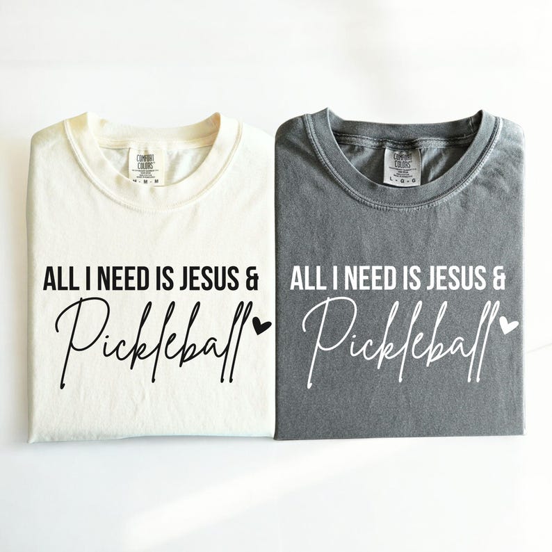 Puede incluir: Dos camisetas con el texto "All I need is Jesus & Pickleball" impreso en ellas. Una camiseta es blanca y la otra es gris.