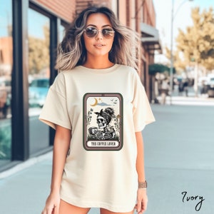 May include: A cream coloured t-shirt with a graphic of a skeleton drinking coffee. The graphic is in a tarot card style with the text "The Coffee Lover".