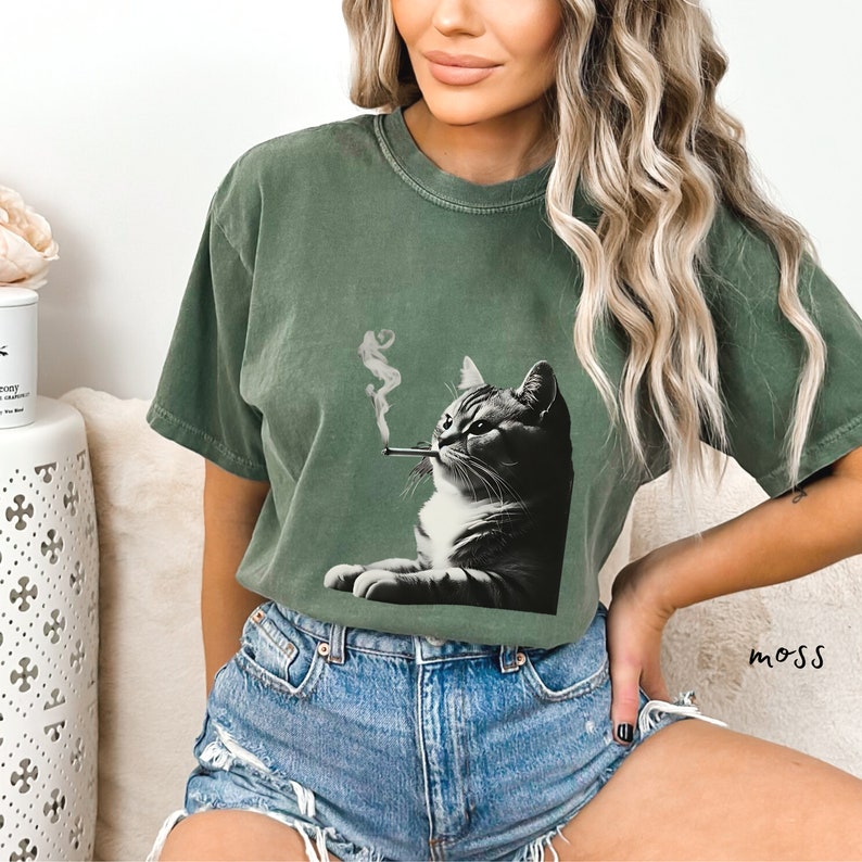 Pu&ograve; includere: Una t-shirt a maniche corte color verde muschio con una stampa grafica di un gatto bianco e nero che fuma una sigaretta con fumo visibile. La maglietta ha un girocollo e una vestibilit&agrave; rilassata, abbinata a pantaloncini di jeans blu sfrangiati.