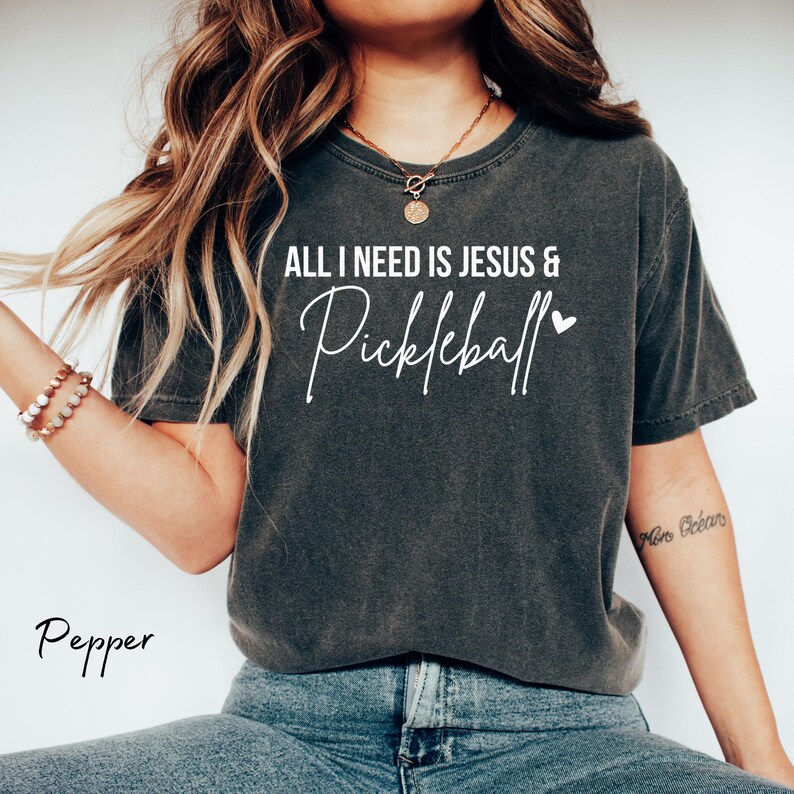 Puede incluir: Una mujer lleva una camiseta gris oscuro con el texto "All I need is Jesus & Pickleball" en blanco. La camiseta tiene un aspecto desgastado y el texto est&aacute; escrito a mano. La mujer lleva pantalones vaqueros azules y tiene el pelo largo y casta&ntilde;o.