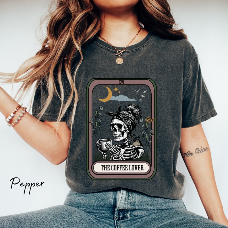May include: A dark grey t-shirt featuring a graphic of a skeleton holding a cup of coffee. The graphic is in a tarot card style with the text "The Coffee Lover" at the bottom. The card is surrounded by a pink border.