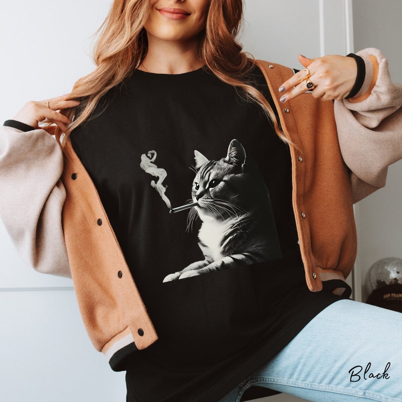 Pu&ograve; includere: Una t-shirt nera con una stampa grafica di un gatto con una sigaretta in bocca, da cui si alza del fumo bianco. Il gatto &egrave; raffigurato in uno stile in bianco e nero, mostrando dettagli della pelliccia e dei baffi sul tessuto nero.