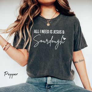 以下が含まれることがあります： ALL I NEED IS JESUS & Sourdoughという白い文字と小さなハートのアイコンがプリントされた、チャコールグレーの半袖Tシャツ。クルーネックで、ヴィンテージ風のウォッシュ加工が施された生地感。下にPepperという文字がプリントされている。