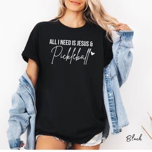 Puede incluir: Camiseta negra con el texto "All I need is Jesus & Pickleball" en letras blancas con un peque&ntilde;o coraz&oacute;n.