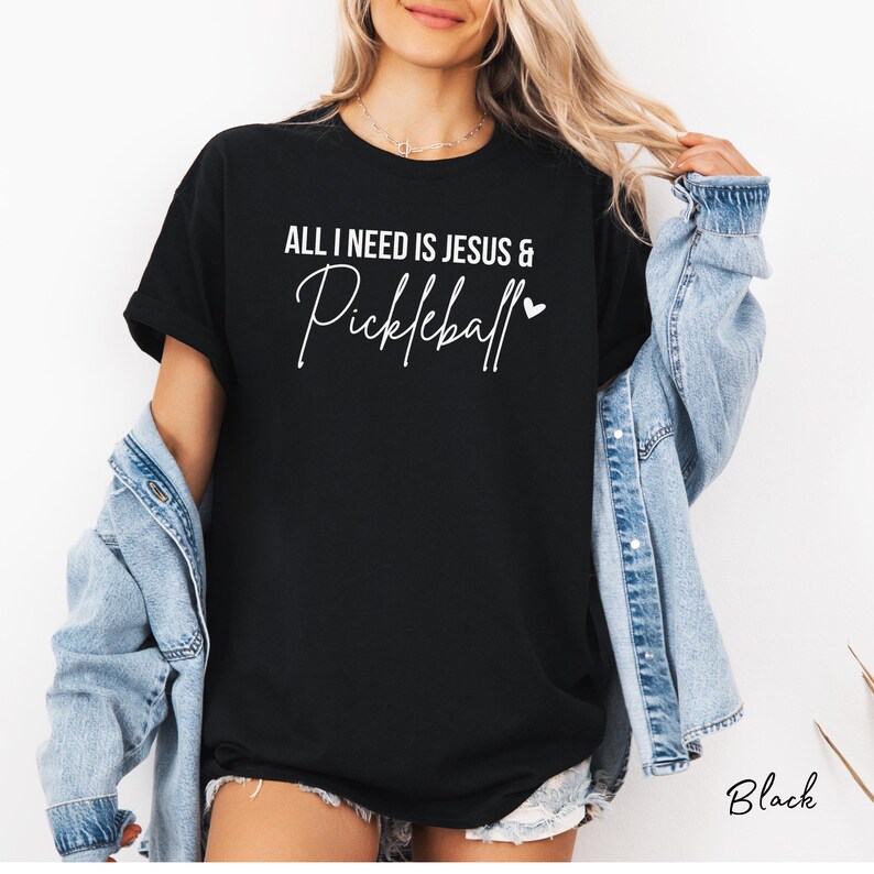 Puede incluir: Camiseta negra con el texto "All I need is Jesus & Pickleball" en letras blancas con un peque&ntilde;o coraz&oacute;n.