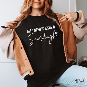 以下が含まれることがあります： 「ALL I NEED IS JESUS & Sourdough」という白い文字と小さなハートのアイコンがプリントされた黒いTシャツ。ベージュとライトブラウンのバーシティジャケットの下に着用されており、裾からはライトブルーのデニムパンツが見えています。