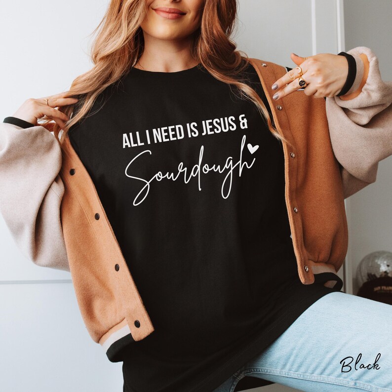 以下が含まれることがあります： 「ALL I NEED IS JESUS & Sourdough」という白い文字と小さなハートのアイコンがプリントされた黒いTシャツ。ベージュとライトブラウンのバーシティジャケットの下に着用されており、裾からはライトブルーのデニムパンツが見えています。